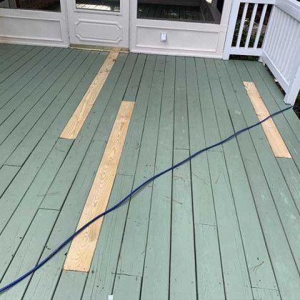Green deck with several tan painted planks and a black cable across it, near white doors and railing.