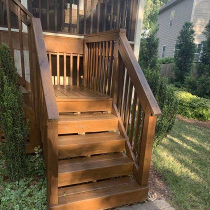 Wooden outdoor stairs with railings leading up to a backyard deck, bordered by grass and shrubs.