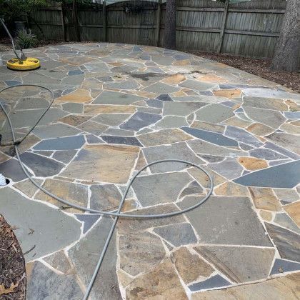 Stone patio in a fenced backyard with a coiled garden hose and a yellow pressure washer.
