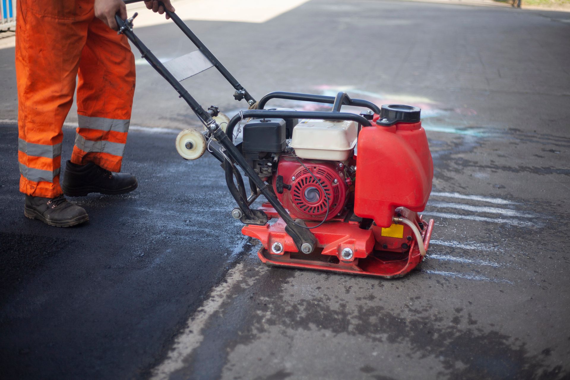 Un trabajador utiliza una apisonadora roja para compactar asfalto negro recién aplicado en una calle.