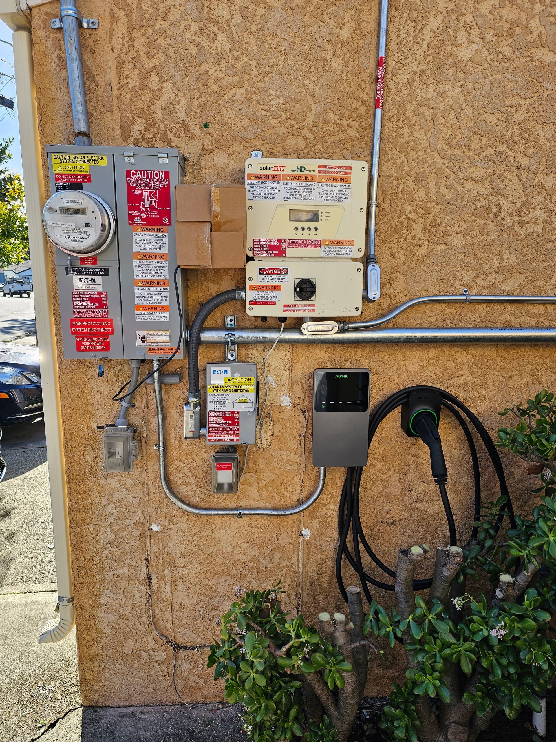 Electrical meter, solar inverter, and EV charger mounted on a stucco building exterior.