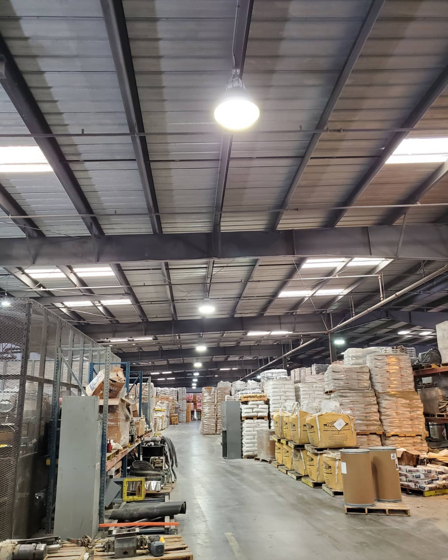 Warehouse interior with rows of pallets, goods, and overhead lighting. Gray metal roof and support beams.