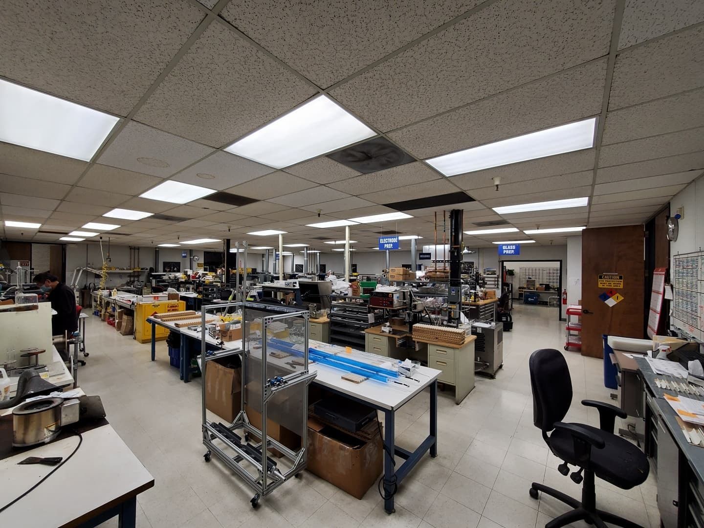 Assembly workshop with workstations, equipment, and people. Fluorescent lighting illuminates the space.
