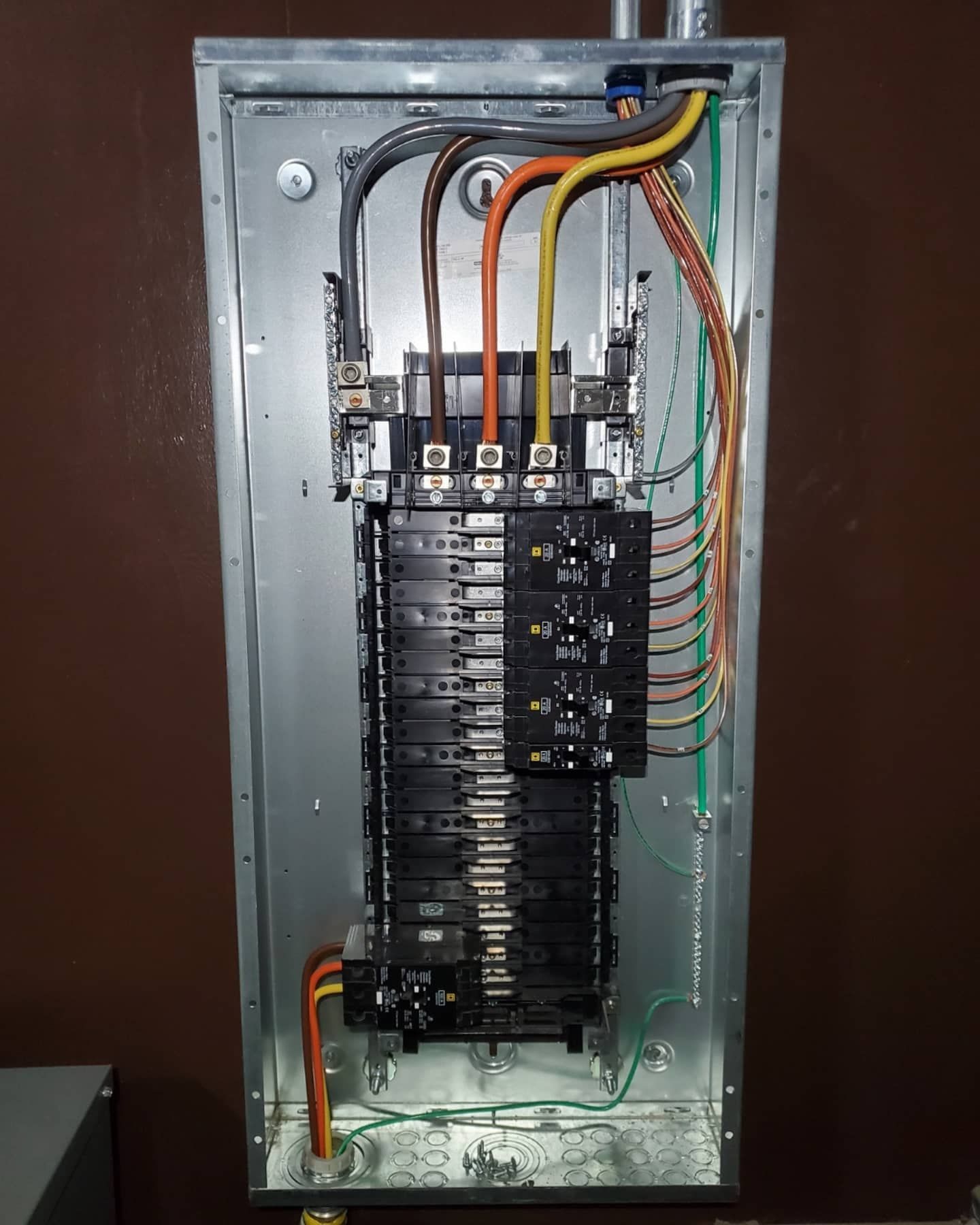 Electrical panel with black circuit breakers and colored wires. Silver metal panel is mounted on a brown wall.