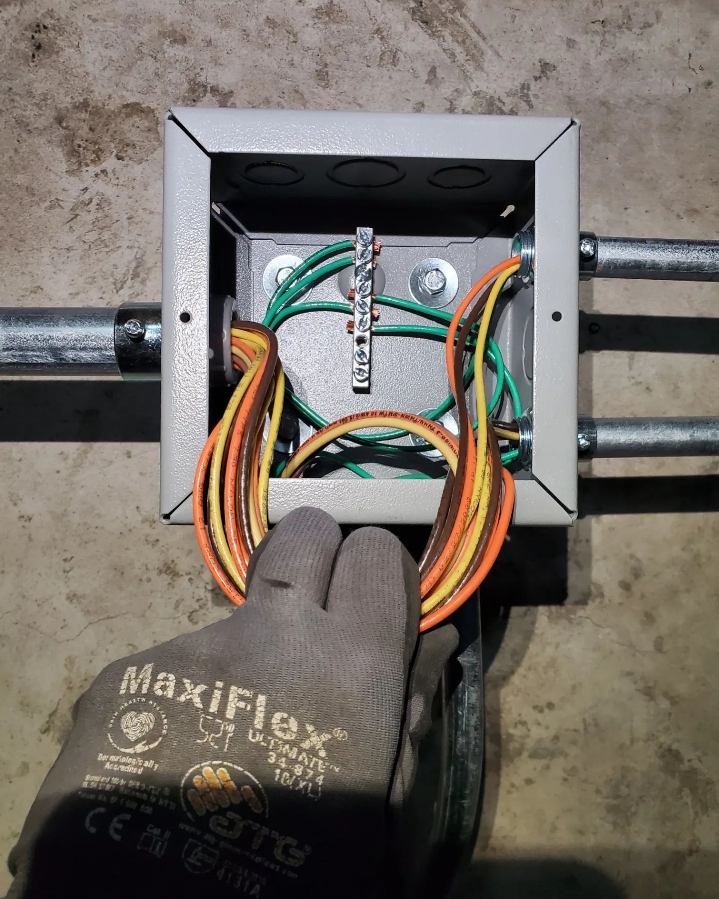 Gloved hand holding wires near an electrical junction box with green and orange wires, and conduit pipes.