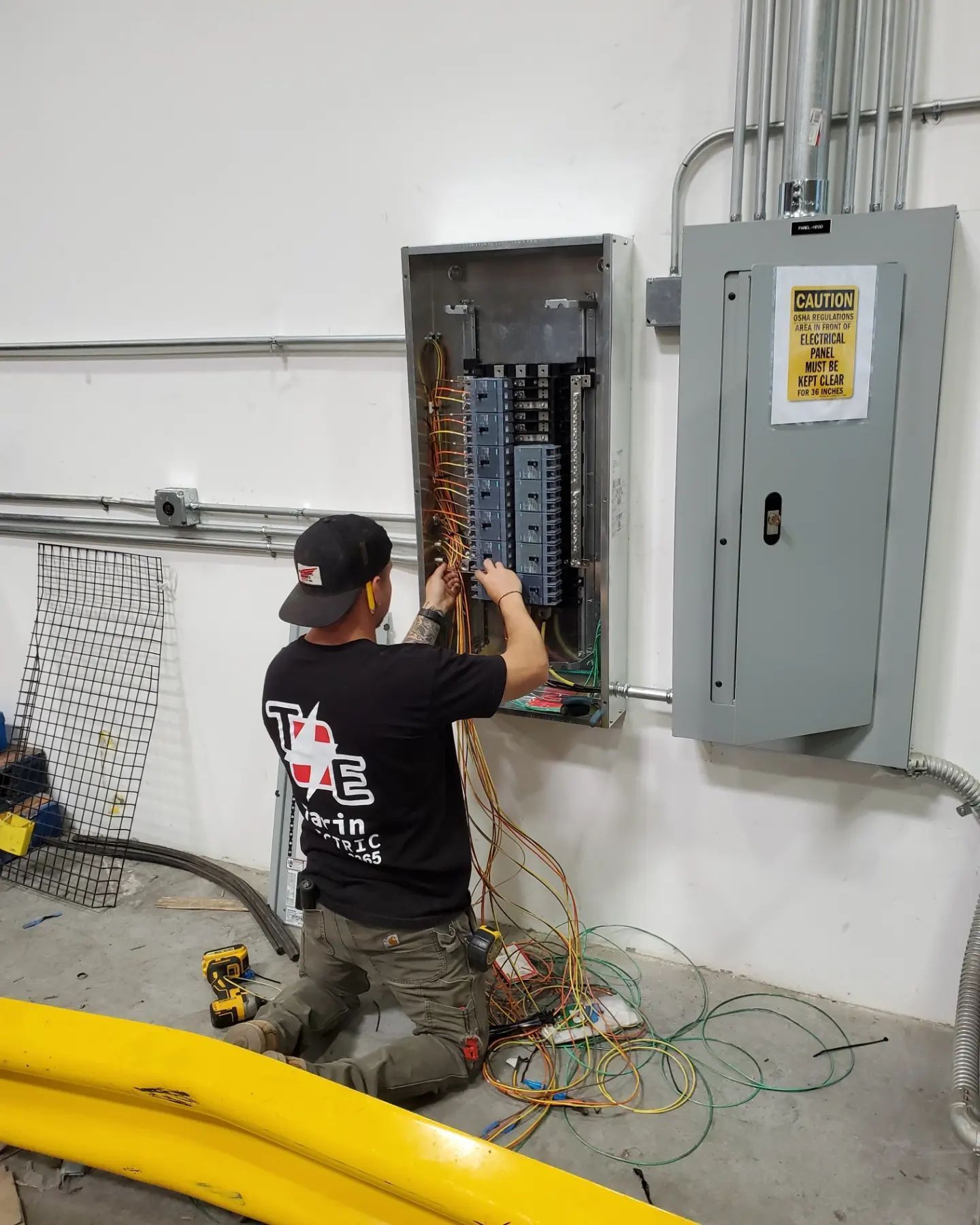 Electrician working on electrical panel, kneeling, with wires and tools in a warehouse setting.