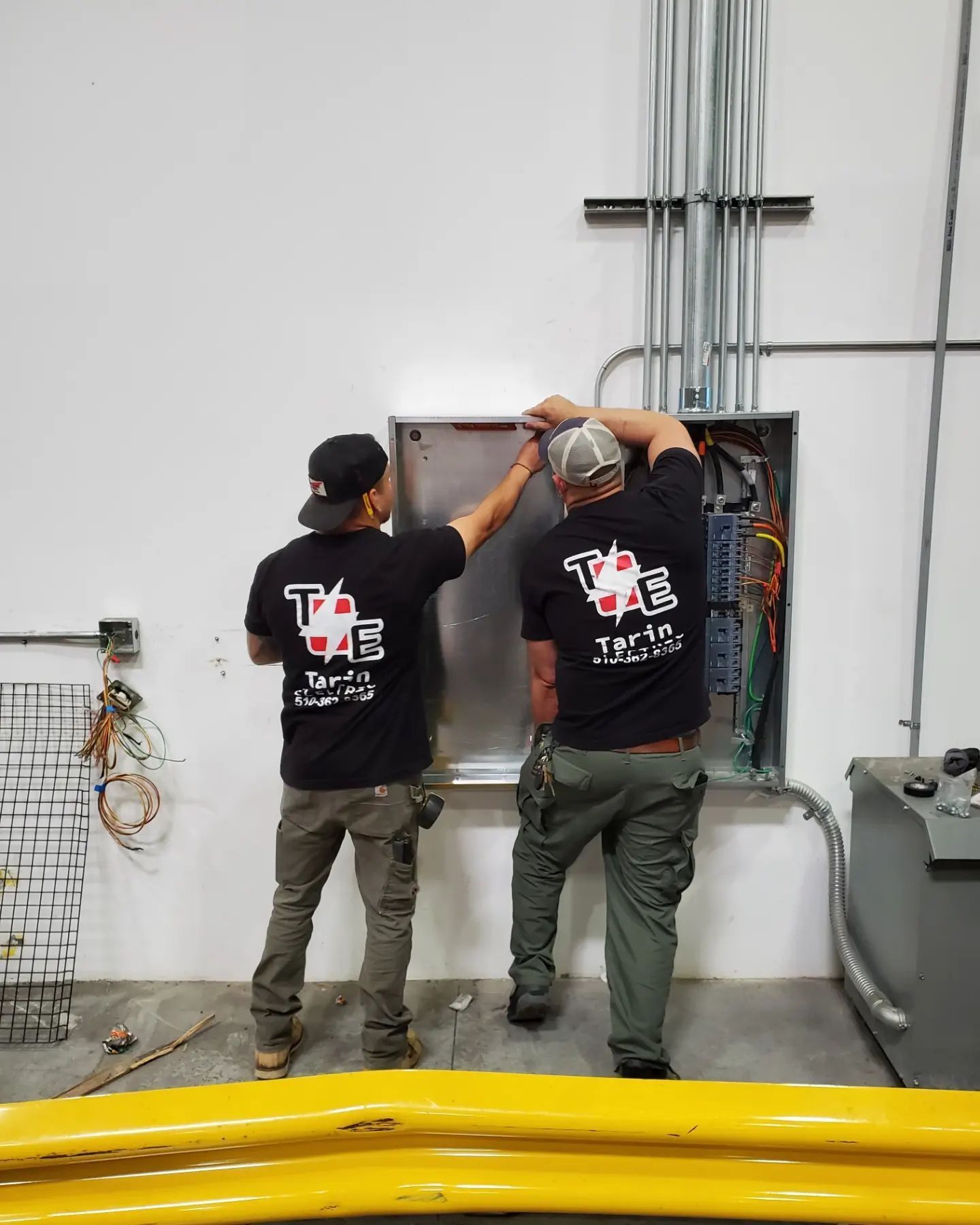 Two electricians working on an electrical panel in a building, wearing black shirts with logo and khaki pants.