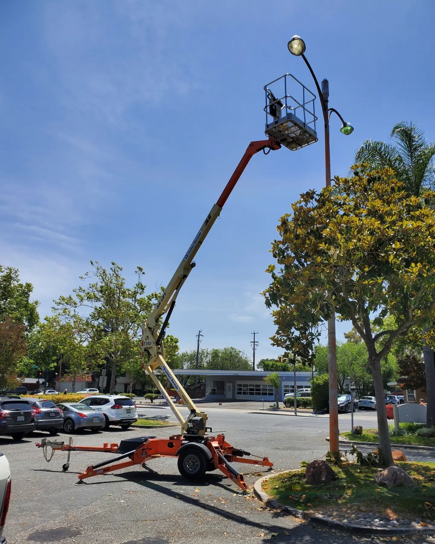 Person in lift working on a light pole in a parking lot. Blue sky, cars, and trees in the background.