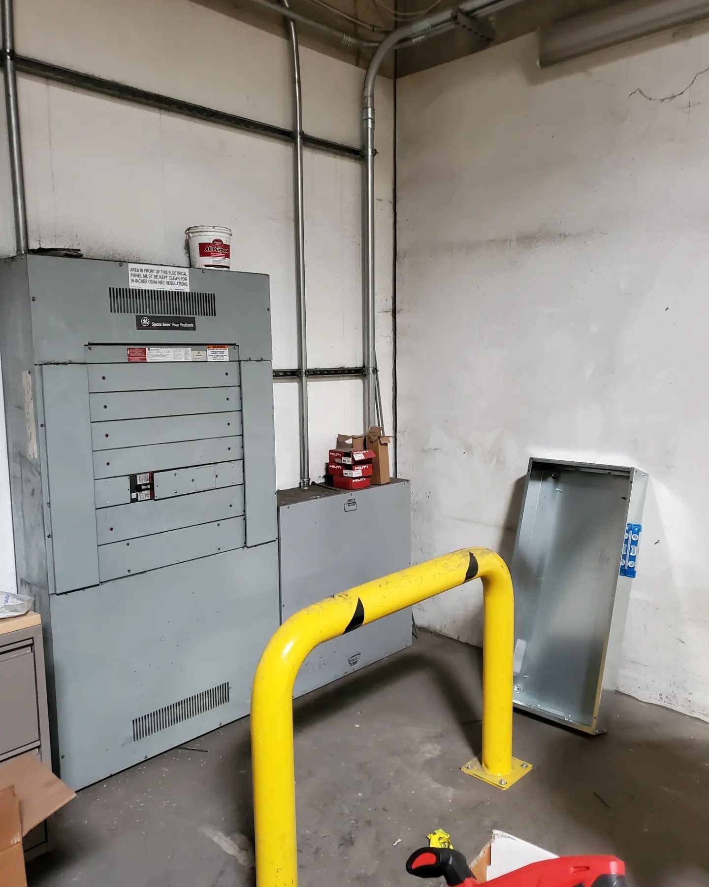 Electrical panel in a room, with yellow protective guardrail. Conduit pipes run vertically.