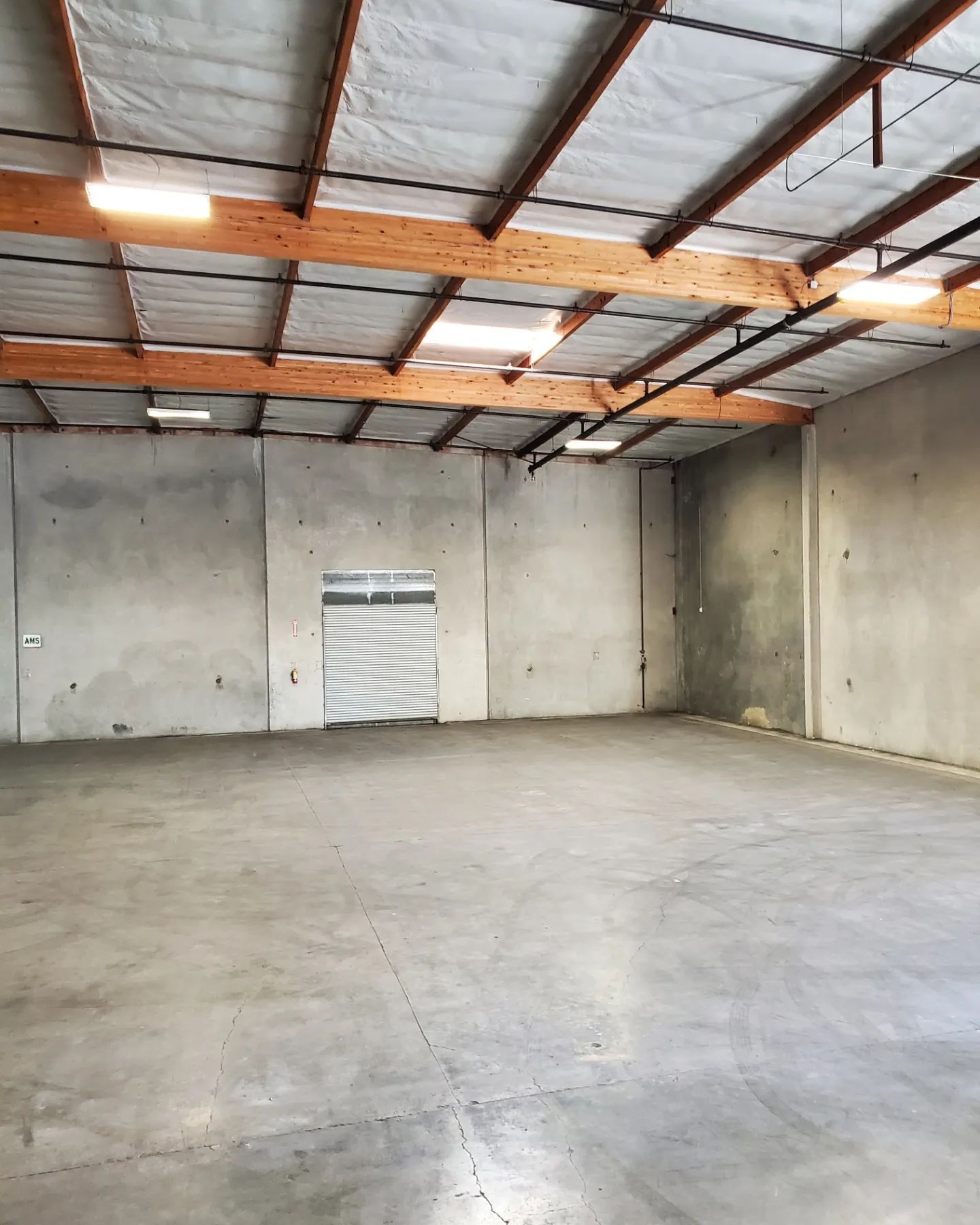 Empty warehouse interior with concrete walls, floor, and ceiling. Wooden beams support the roof with recessed lights.