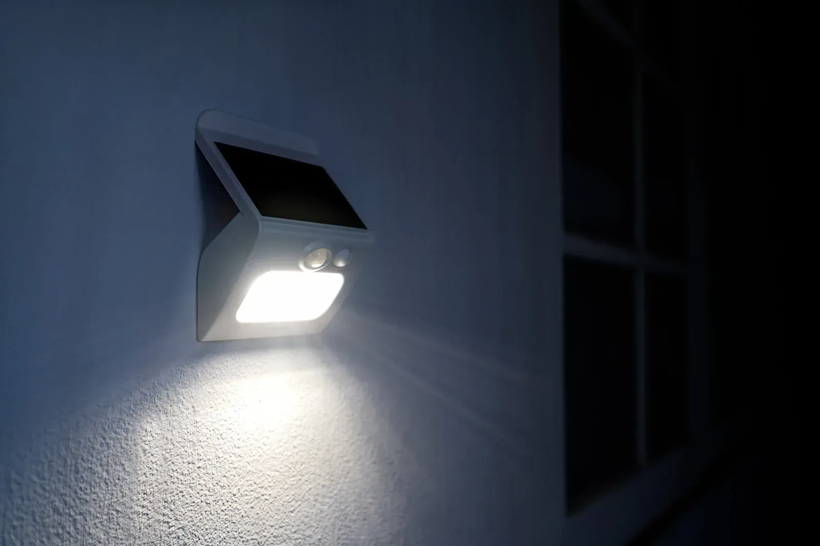 White solar-powered outdoor light fixture on a wall; it is illuminated at night, casting light below.