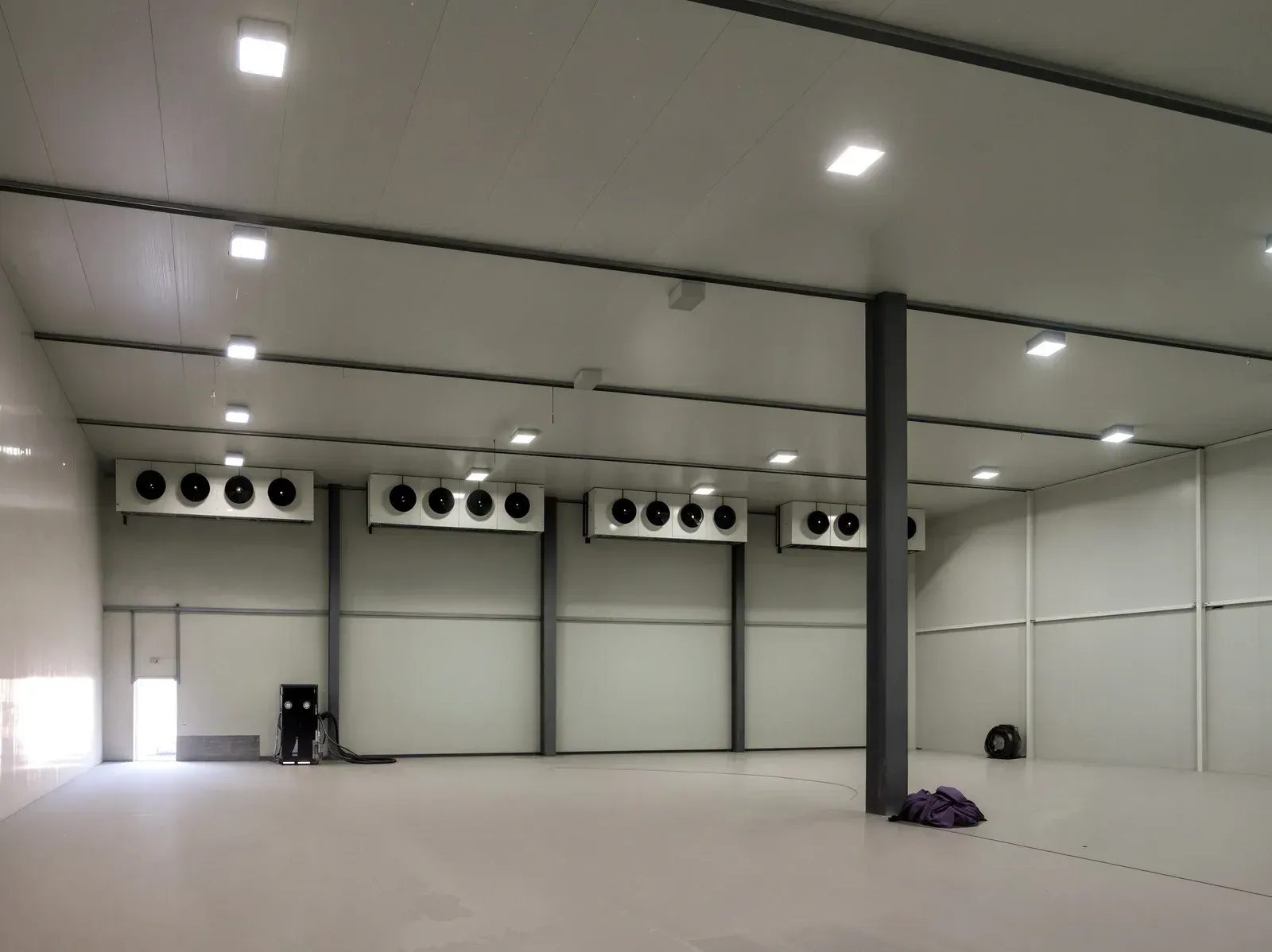 Large, empty, white-walled cold storage room with ceiling lights, cooling units, and a central support column.
