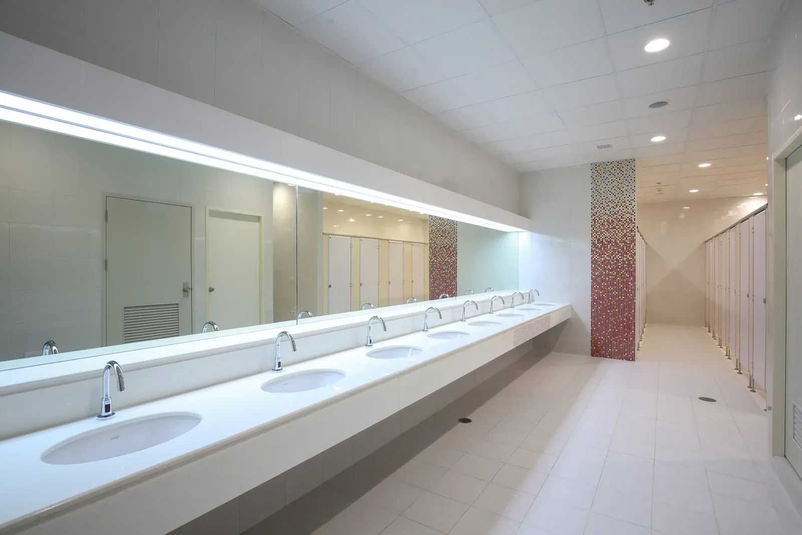 Long, bright public restroom with sinks, mirrors, and stalls. White and minimalist design.