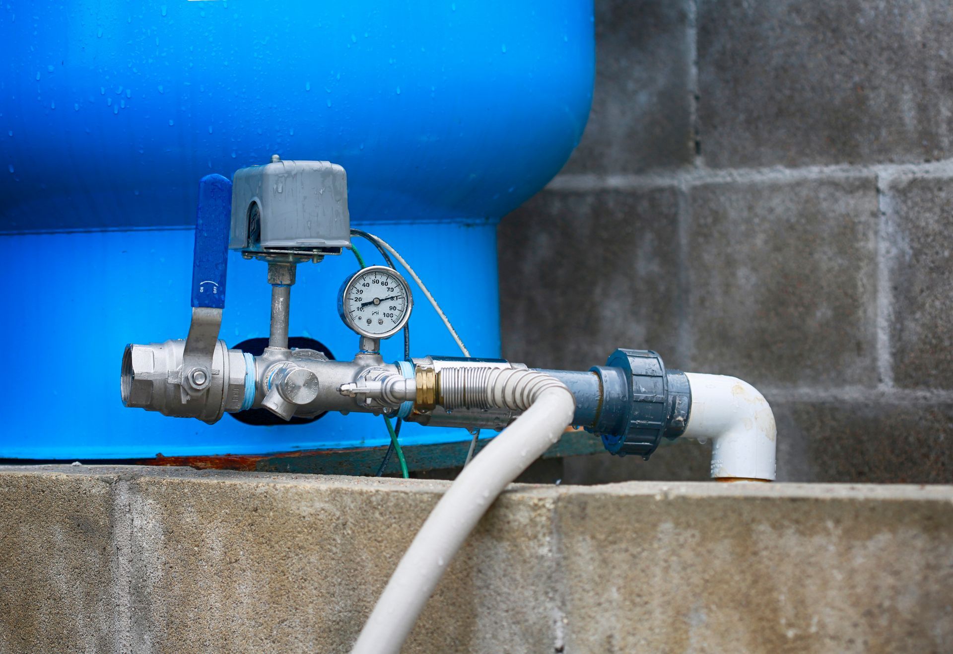 Blue water tank with attached pipes, gauge, and electrical box; outdoors on a concrete base.