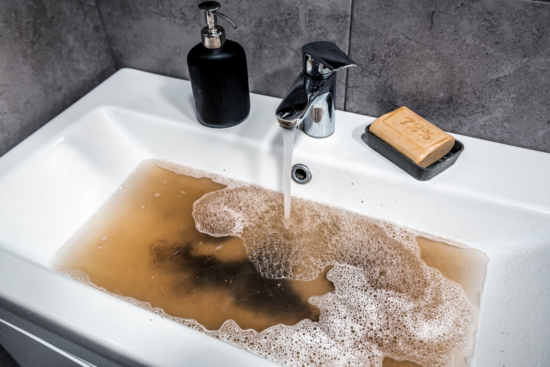 Brown, murky water fills a white bathroom sink as tap water runs. A soap dish and dispenser sit nearby.