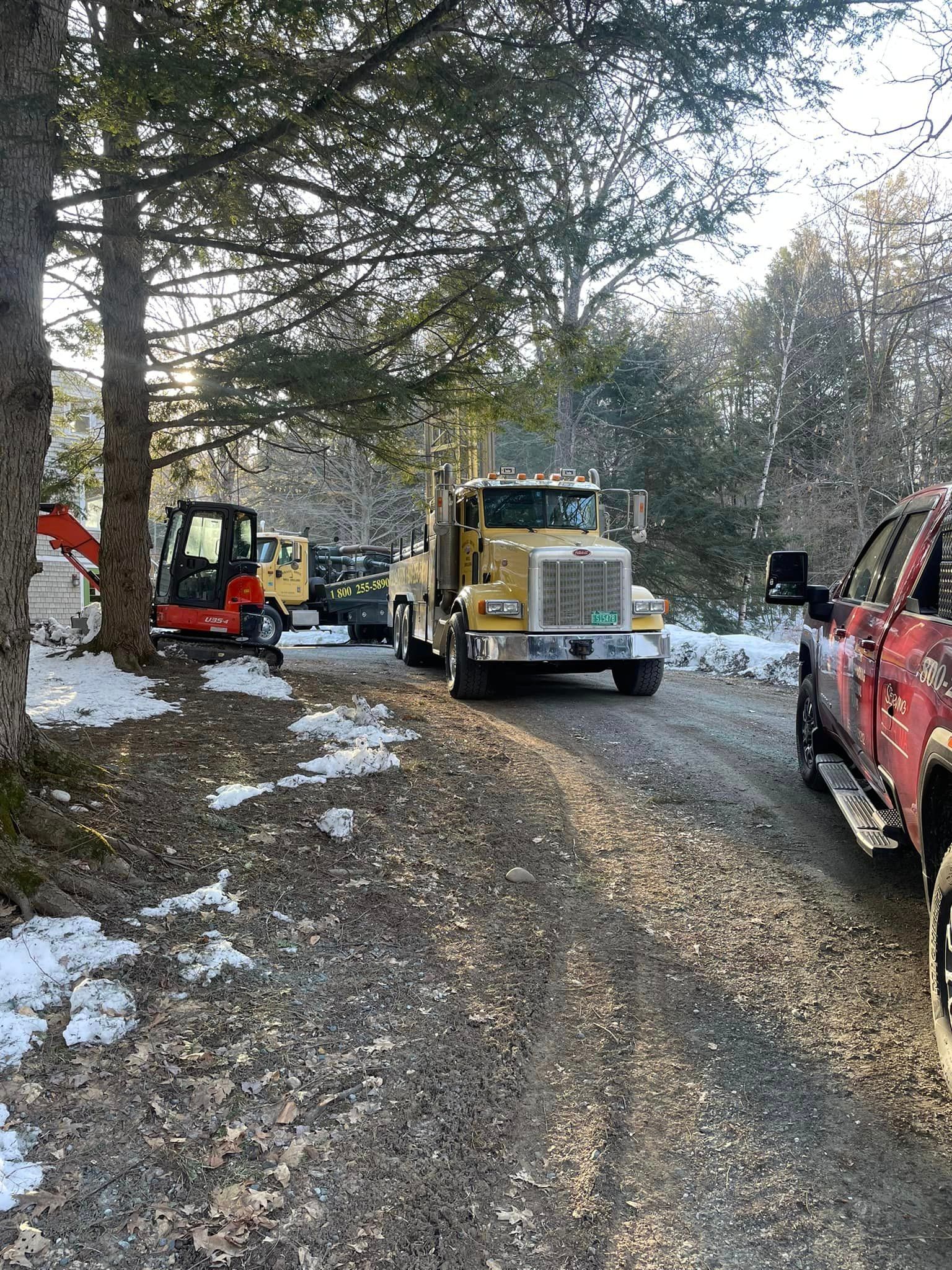 Yellow utility truck and small excavator on a snowy gravel road beside trees, with a red truck parked nearby.