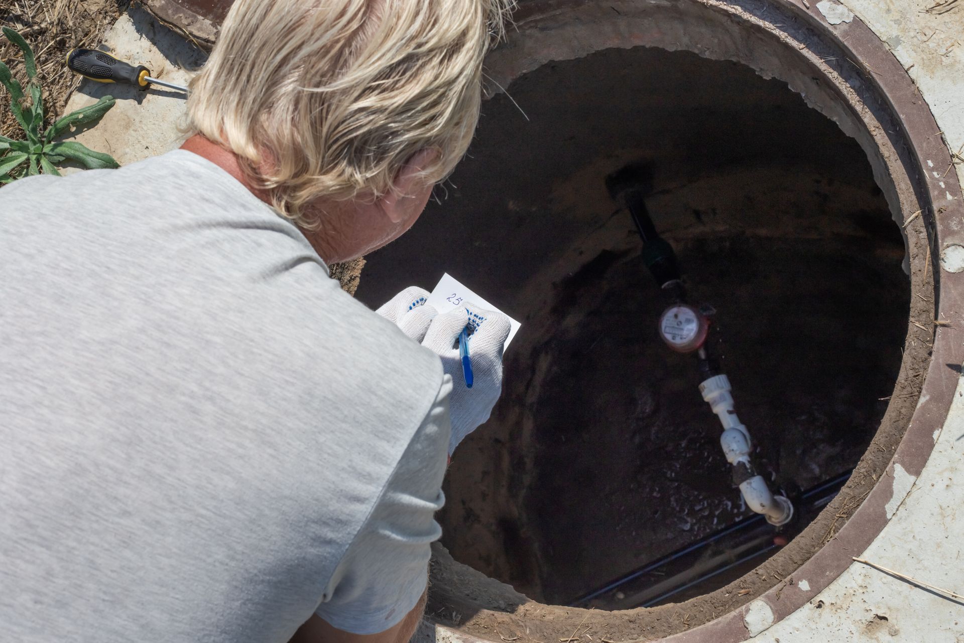 A man is looking into a manhole while holding a piece of paper.
