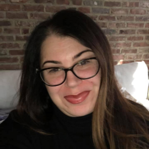 A woman wearing glasses is smiling in front of a brick wall