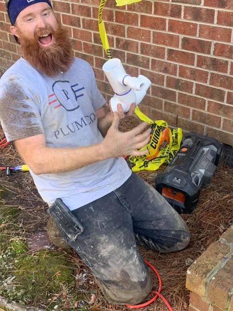 Plumber kneeling outdoors by brick wall, holding a white PVC pipe fitting and smiling.