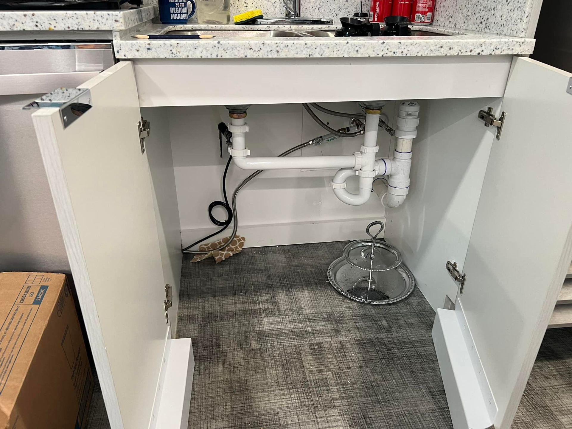 Open cabinet under sink showing plumbing pipes, drain, and a metal bucket on the floor.