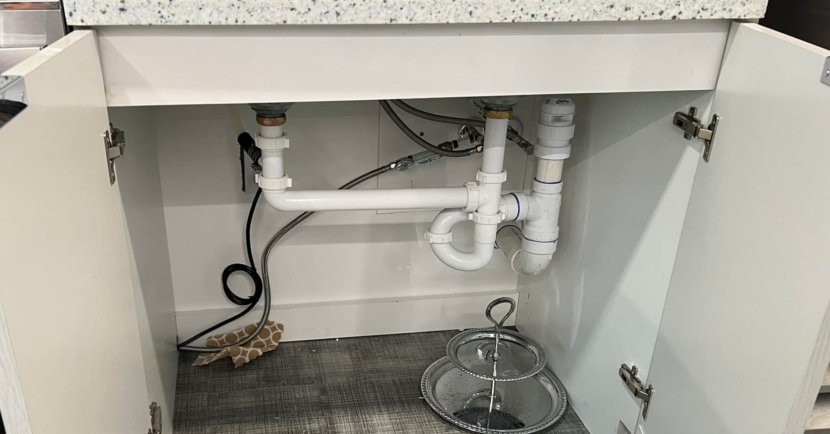 Open cabinet under sink showing white plumbing pipes and a drain trap on a gray countertop.