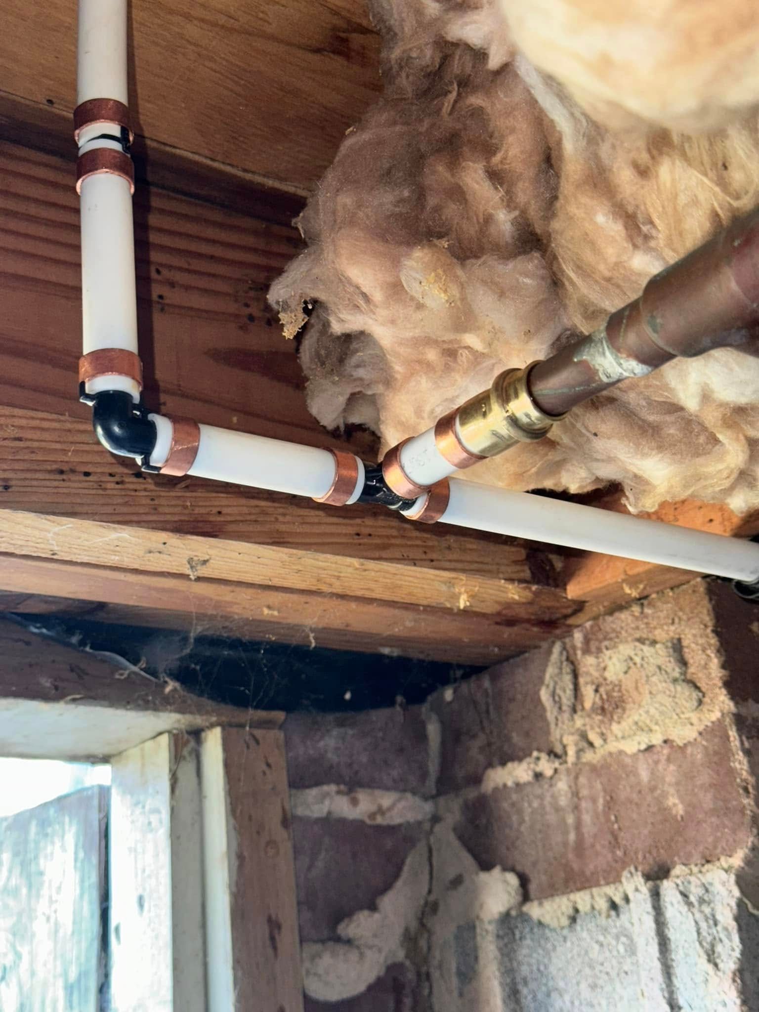 Insulated attic plumbing with white and copper pipes running across exposed wooden beams and rock insulation.