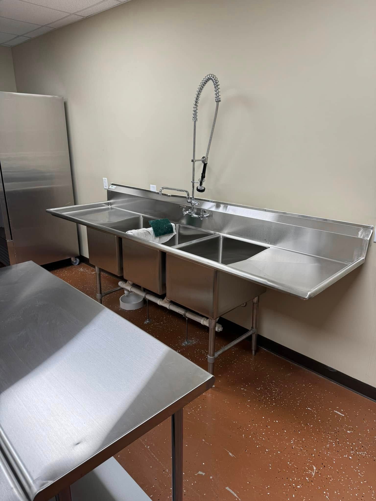 Stainless steel commercial kitchen sink and prep table in a tiled room with speckled floor.