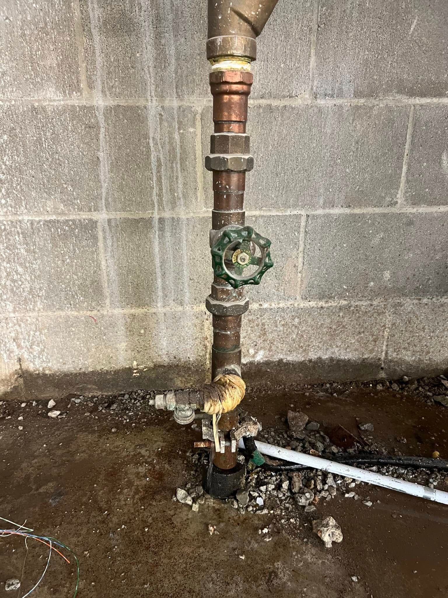 Vertical copper pipe with green valve against a cinder block wall, near a wet floor.