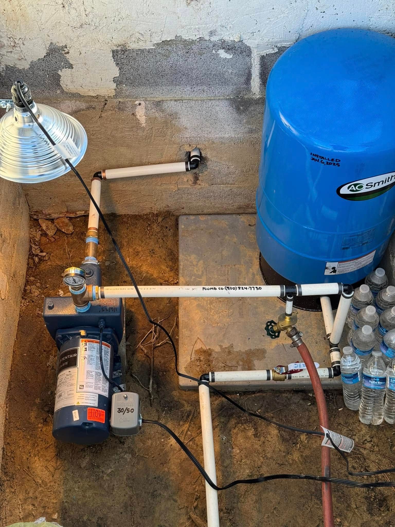 Water heater and pump system with blue tank, pipes, and wiring against a concrete wall.