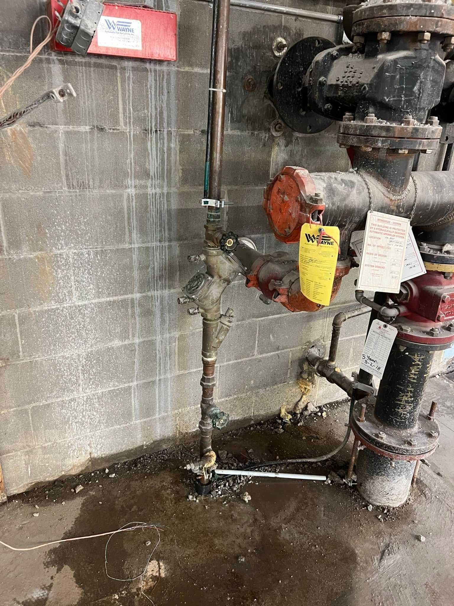 Industrial pipes and valves against a cinderblock wall with a wet concrete floor and a red electrical box.
