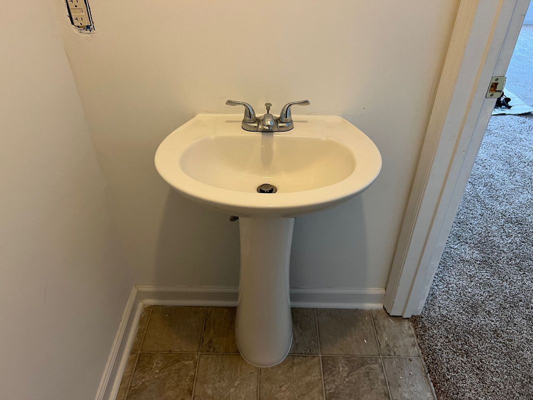 Small white pedestal sink in a tiled bathroom corner with chrome faucet and drain.