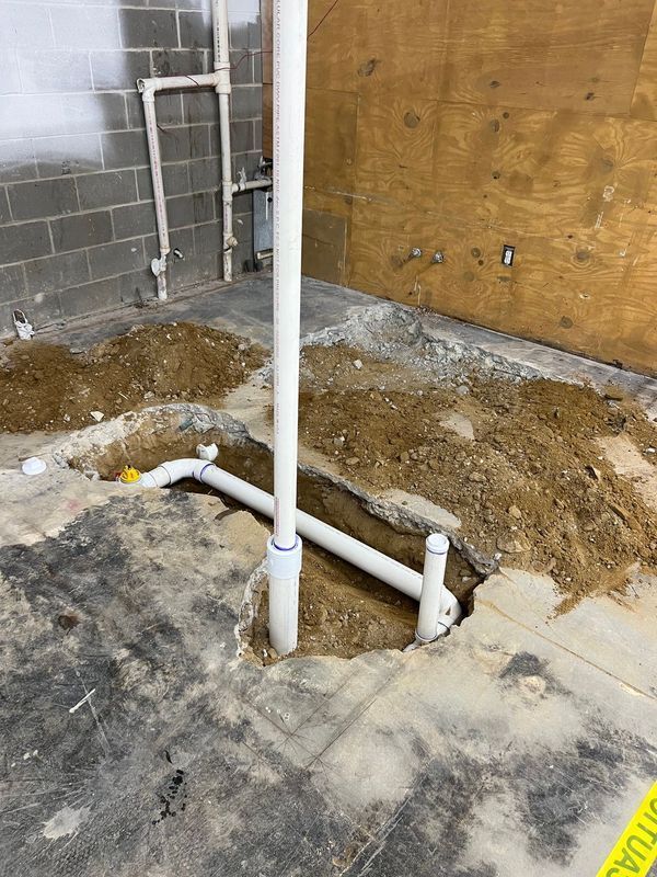 Plumbing trench with exposed white pipes beside a concrete floor and plywood wall