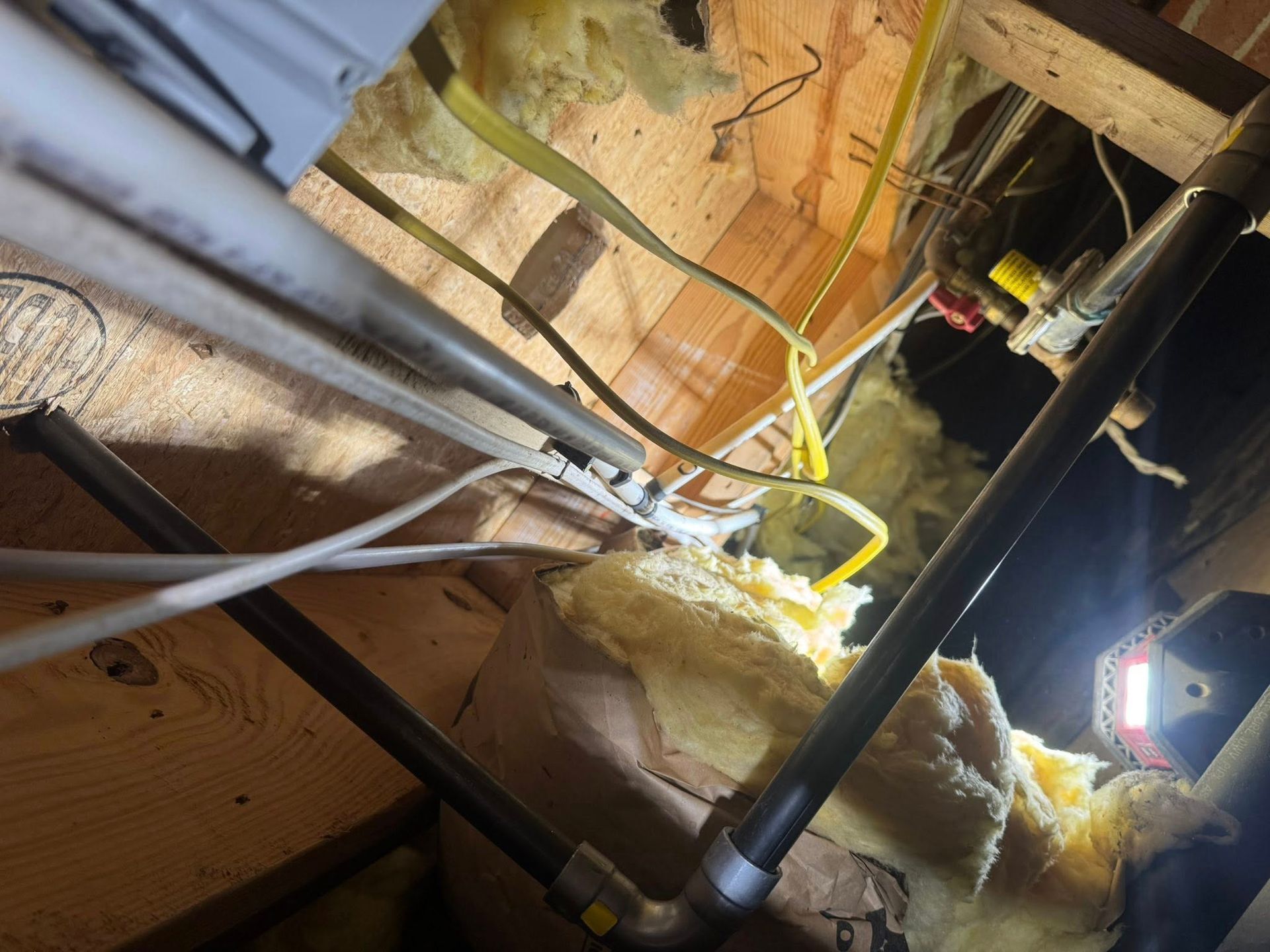 Cluttered attic crawlspace with pipes, wiring, insulation, and a flashlight illuminating the area