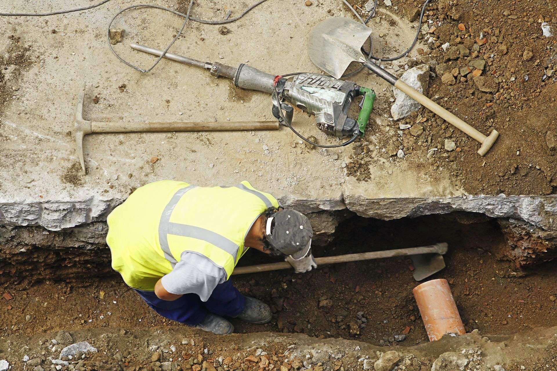 Worker in a neon vest crouches in a trench beside exposed pipes and a jackhammer.