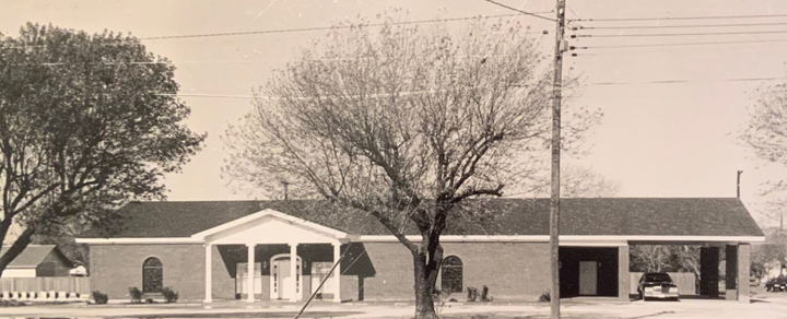 Black-and-white photo of a long, low house with a central porch and trees in front