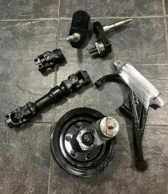 Piezas de dirección y suspensión de metal negro desmontadas, dispuestas sobre un suelo de baldosas grises.
