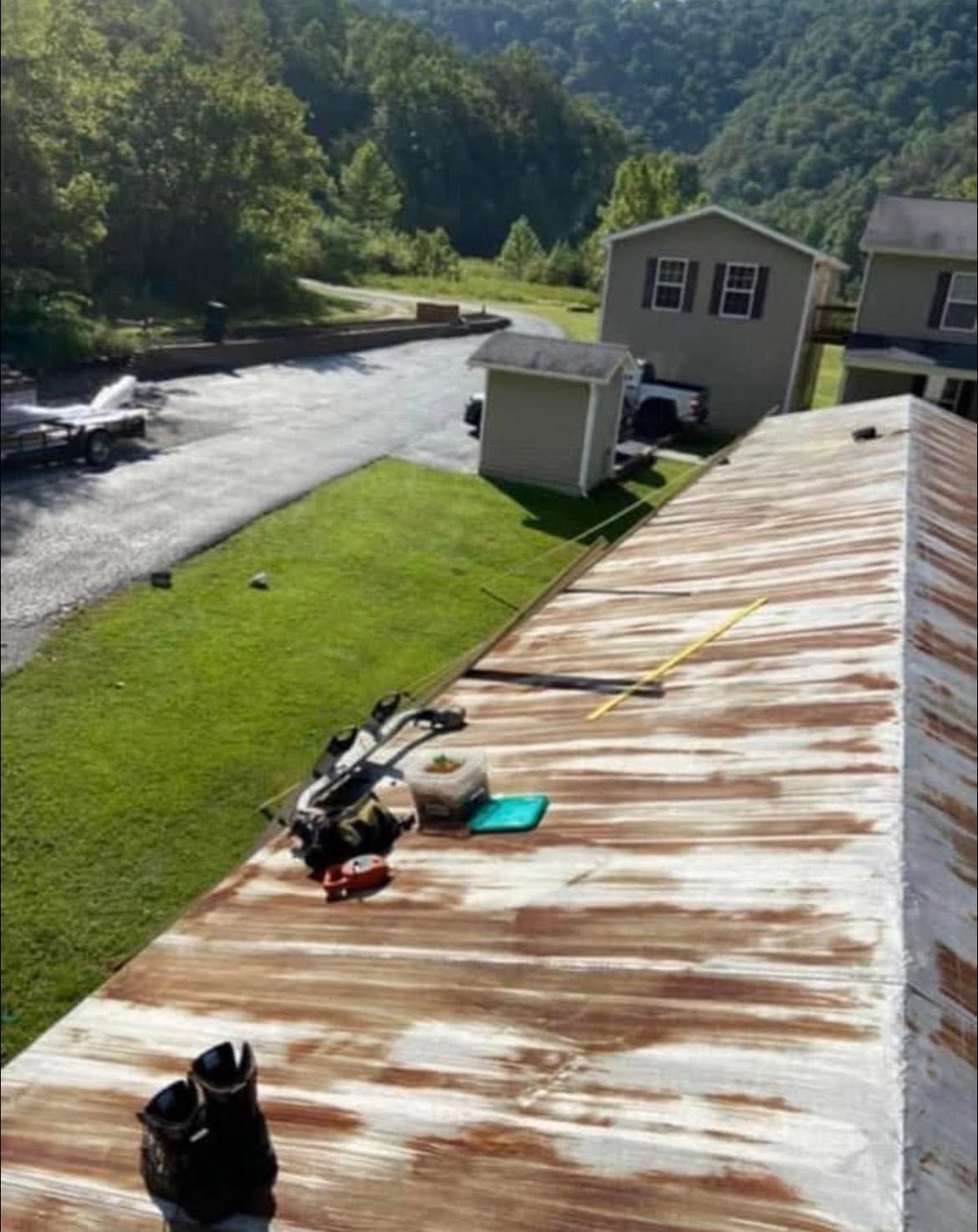 Workers on a rusty metal roof by a river, with trees, a house, and hills in the background.