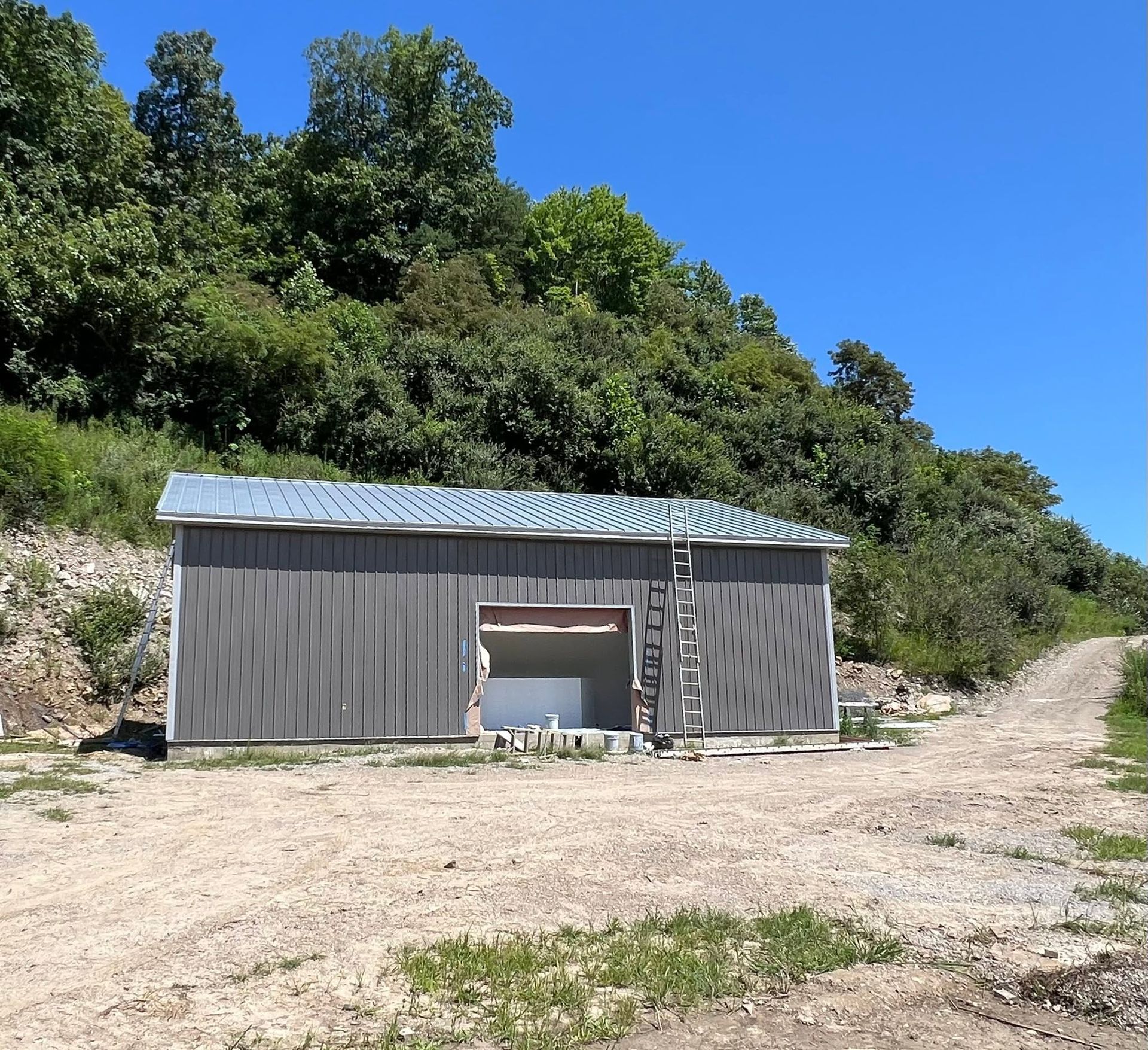 Small gray shed with metal roof beside a dirt road, set against a wooded hillside under a clear blue sky.