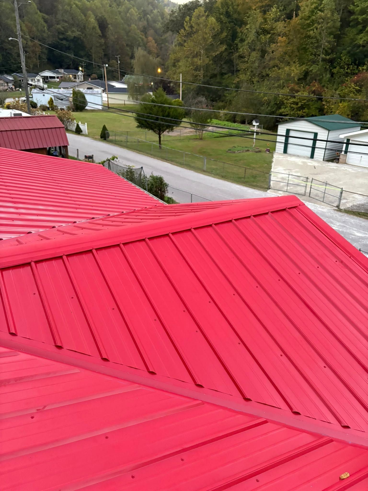 Bright red metal roof in foreground, with a walkway, trees, parked cars, and hillside buildings beyond.