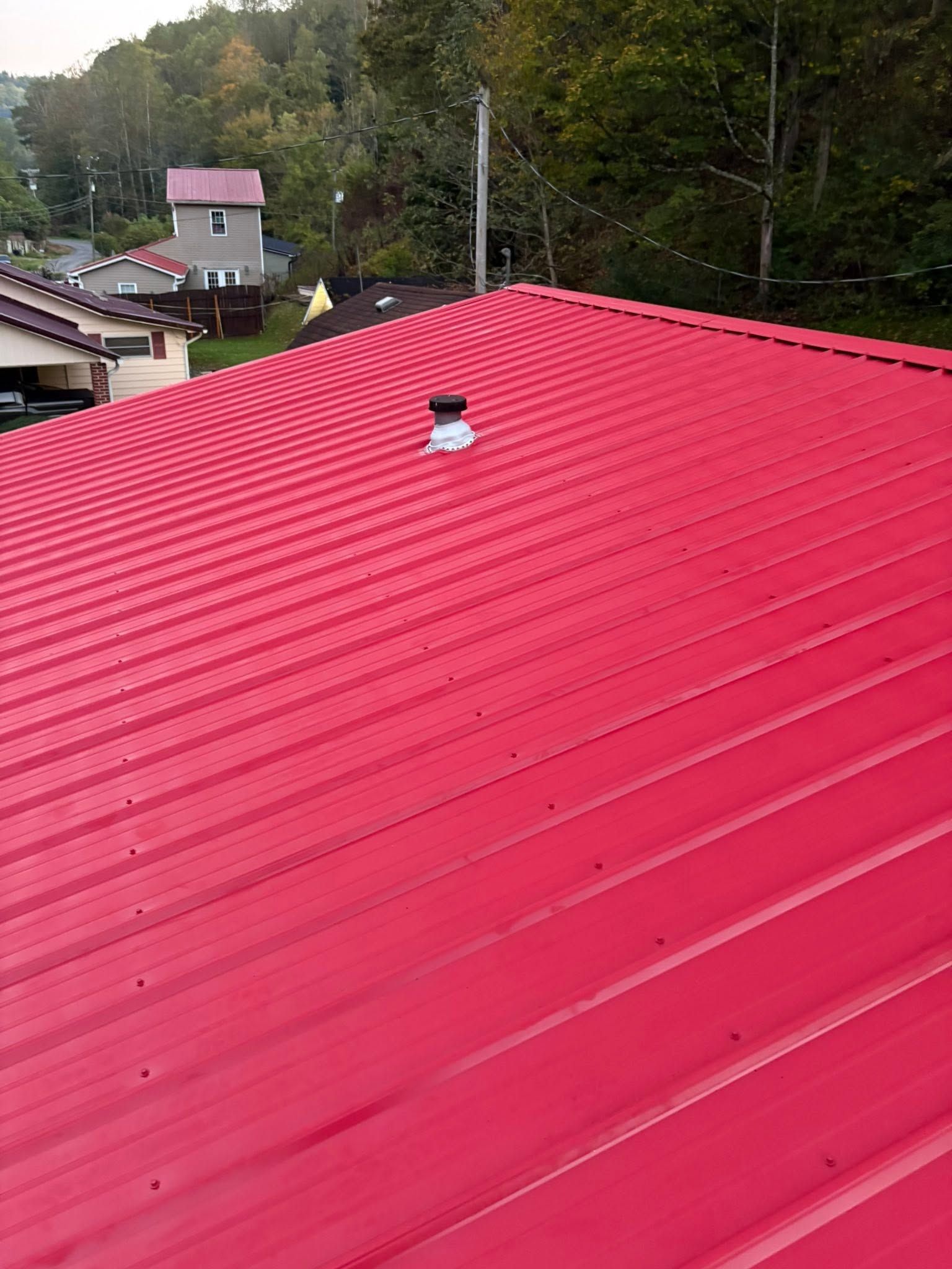 Bright red metal roof with a small vent, viewed over a wooded hillside neighborhood.