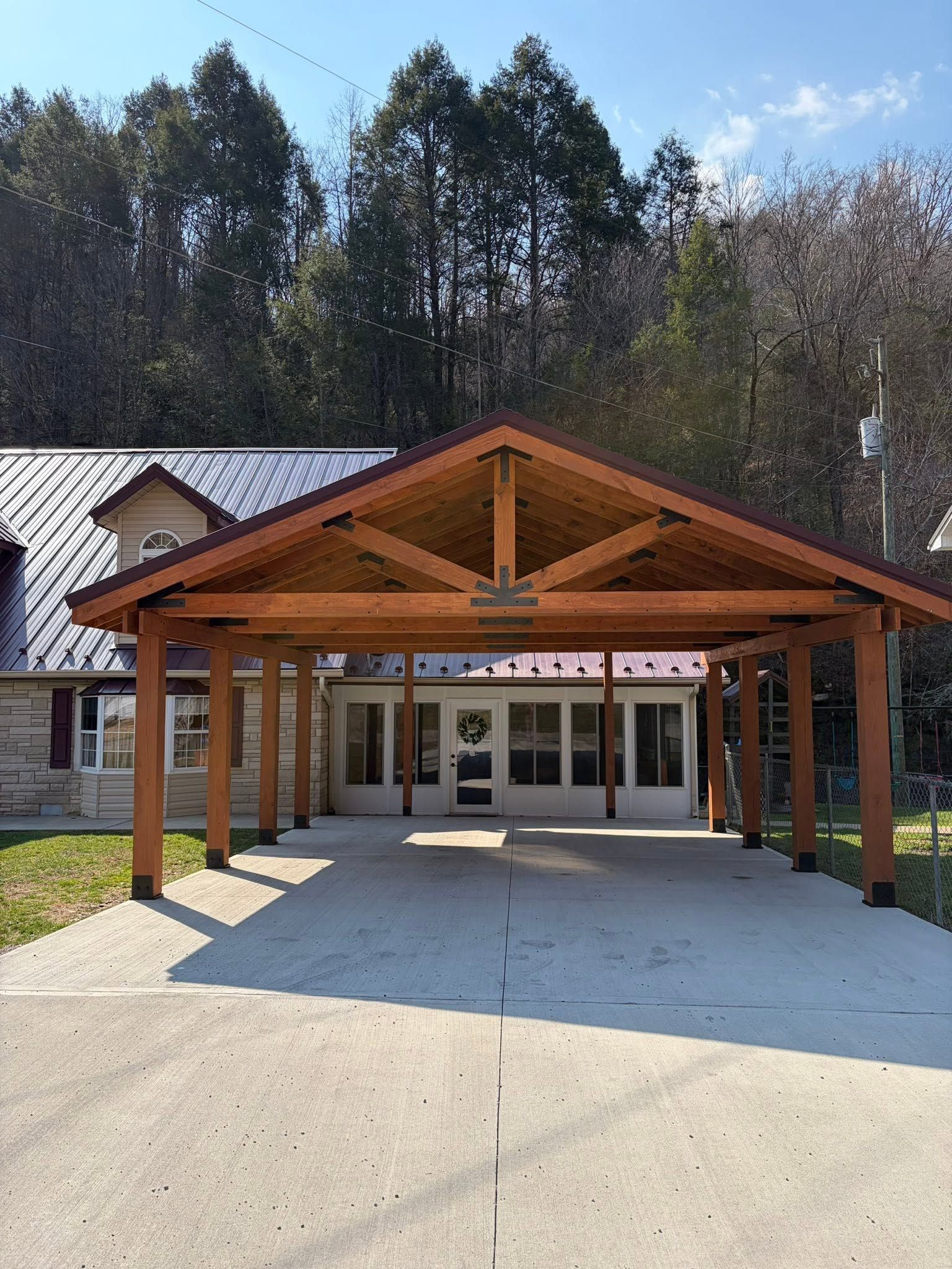 Wooden pavilion entrance with a peaked roof in front of a building, surrounded by trees and sunlight.