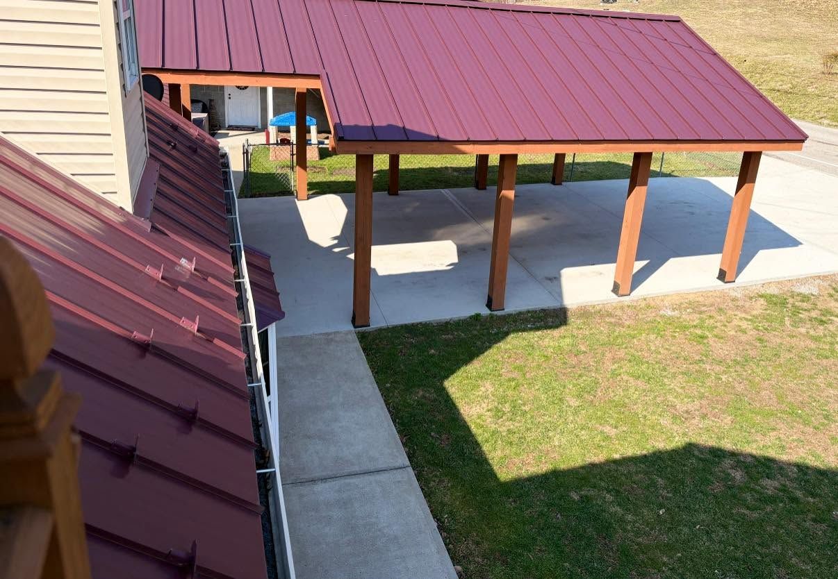 Covered concrete patio walkway beside a house with a red metal roof and grassy yard