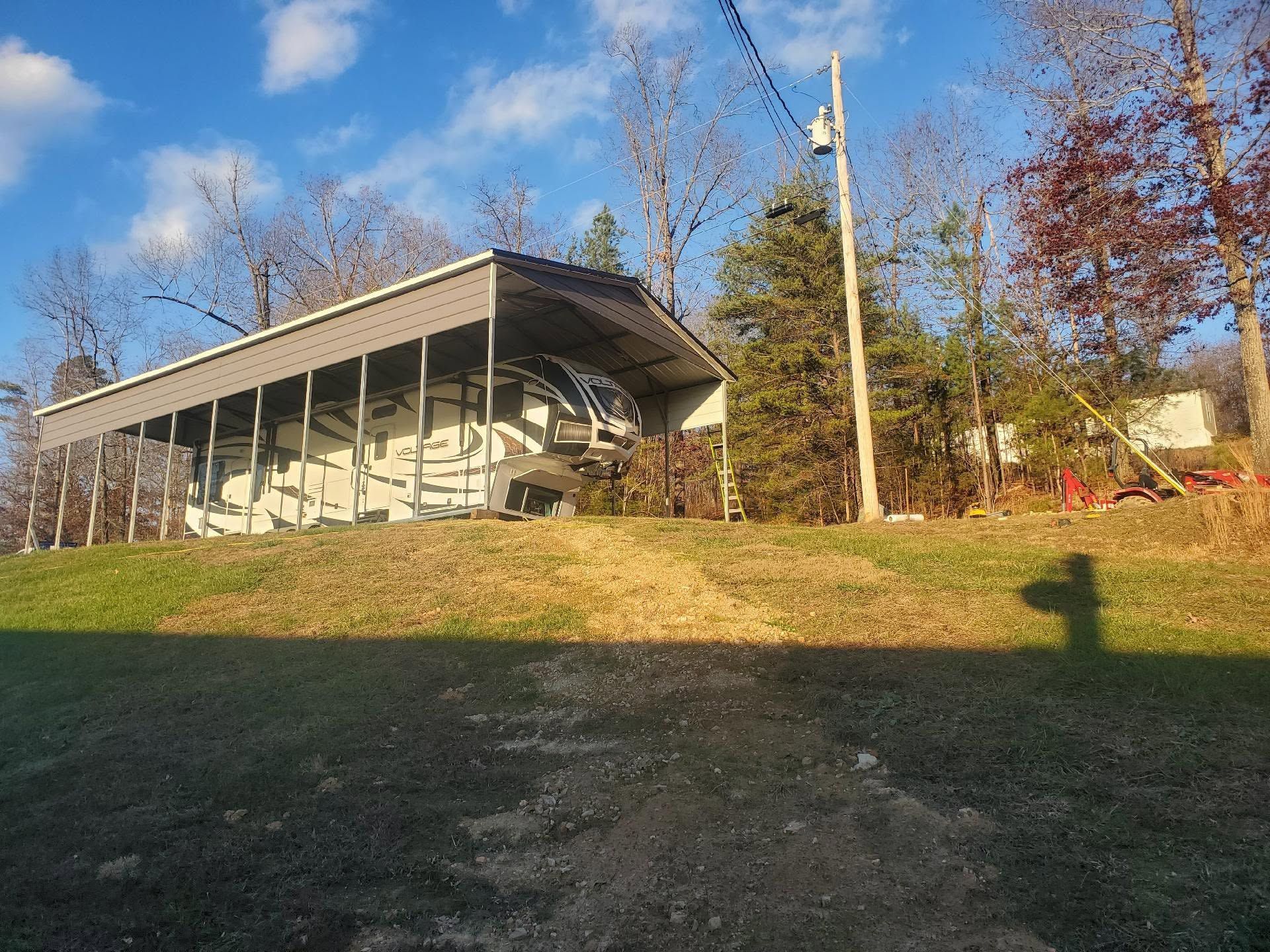 Open-sided pavilion on a grassy hill under a blue sky, with trees and a lamppost nearby