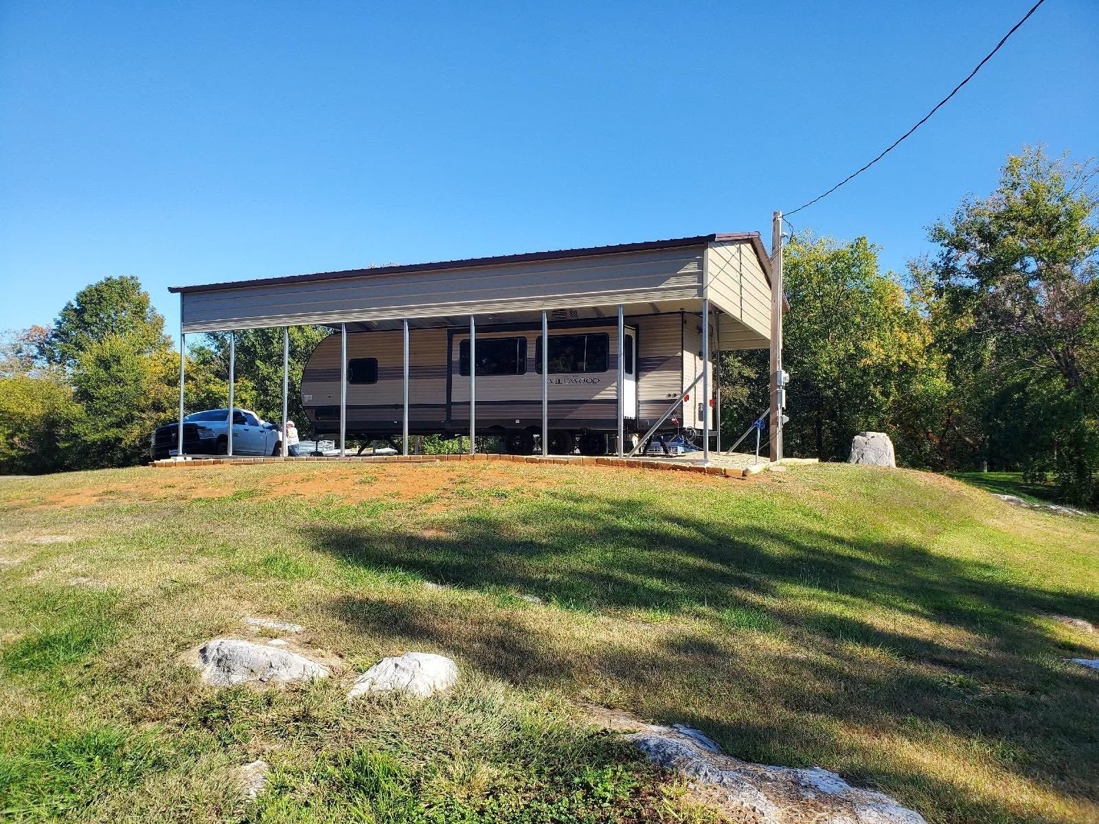 Small cabin on a grassy hill under a clear blue sky, with trees and rocks in the foreground