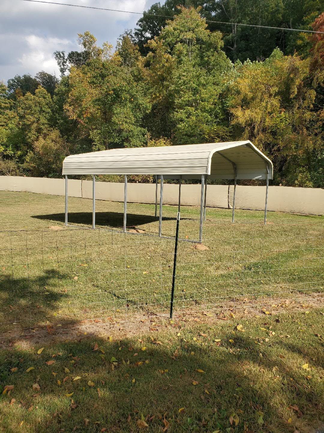 Small metal-framed shelter in a grassy yard with trees and a fence in the background.
