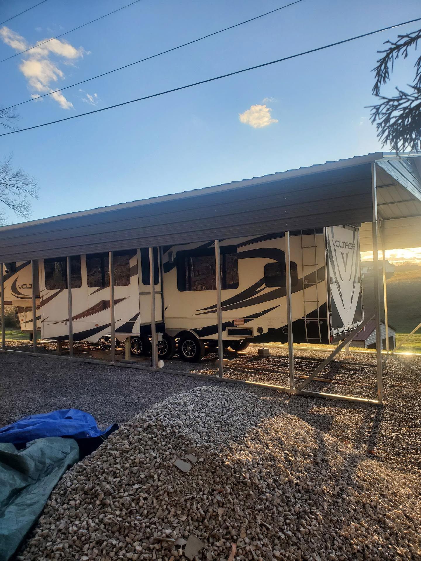 RV under a metal carport on a gravel lot at sunset, with desert hills in the background.