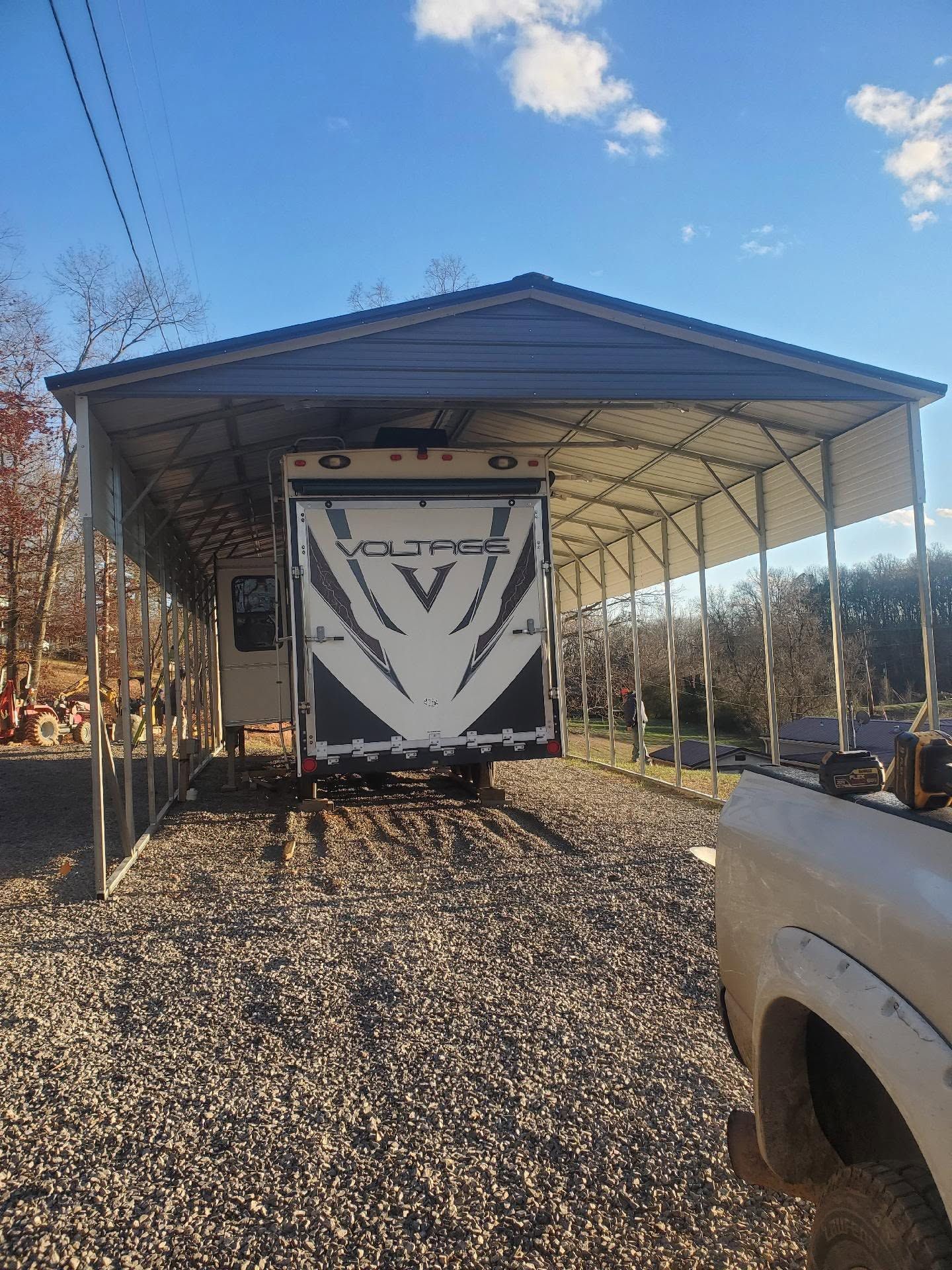 RV under a metal carport on a gravel lot, with a white truck at right and blue sky above