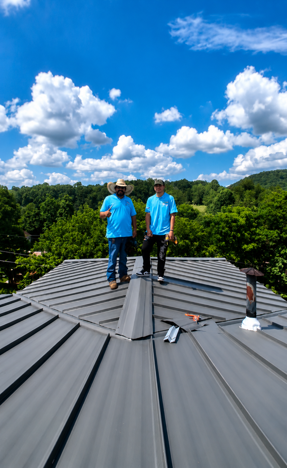 Two workers in blue shirts standing on a gray metal roof under a bright blue sky with clouds.
