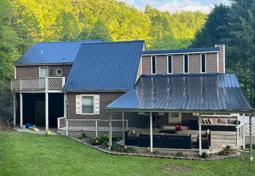 Two-story house with blue metal roofs, porch, and green lawn in a wooded setting