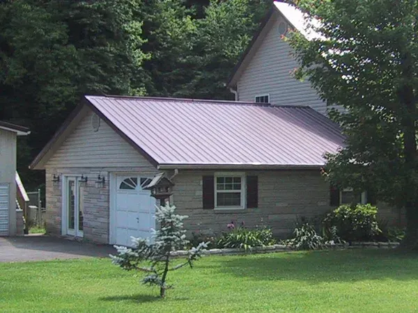 Small house with a purple metal roof, white garage door, and green lawn surrounded by trees.